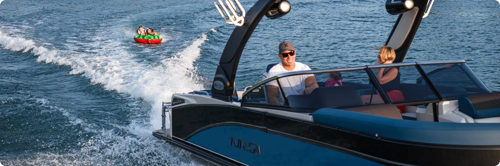 A man and woman enjoying a ride in a motorboat on a sunny day, with two people tubing on an inflatable raft behind them in the water. Waves splash around the boat and raft as they speed across the blue water.