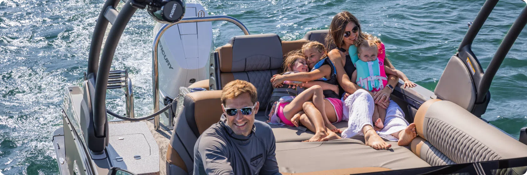 A family enjoys a sunny day on a boat. A man steers while a woman sits with three children, two wearing life jackets, on a cushioned area. The water is calm, and they all appear relaxed and happy in the sun.