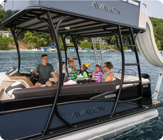 A family enjoys a boat ride on a sunny day. Two children wearing life jackets and party hats sit between two adults. The boat is on a lake surrounded by green trees and houses in the background. The boat has "Avalon" written on it.