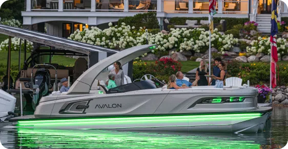 A sleek pontoon boat from leading brands, adorned with green LED lights, floats near a dock beside a house with a garden. Several people stand and sit on the boat, savoring the picturesque scene. Colorful flags flutter on a pole by the dock, adding to the festive ambiance.