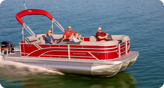 A red pontoon boat with a sunshade canopy, possibly one of the top pontoon boat brands, is cruising on a calm lake. Four people are seated comfortably, enjoying the ride under clear skies. The water around them is a deep blue-green.