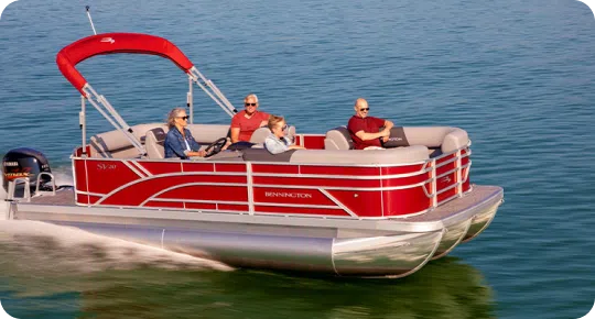 A red pontoon boat with a sunshade canopy, possibly one of the top pontoon boat brands, is cruising on a calm lake. Four people are seated comfortably, enjoying the ride under clear skies. The water around them is a deep blue-green.