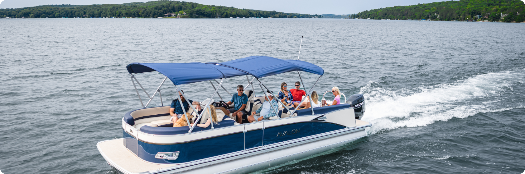 A group of people are relaxing on a luxury pontoon boat with a canopy, cruising on a wide lake. Known for top pontoon boat brands, they enjoy the tranquil ride as green, forested hills line the shore in the background under a clear sky.