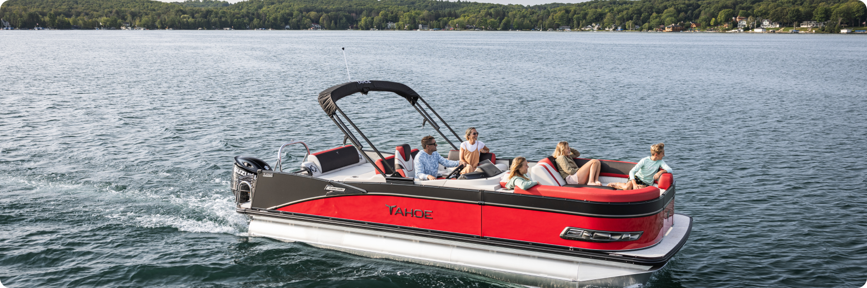 A group of people relaxes on a red and black Tahoe pontoon boat, one of the top pontoon boat brands, cruising on a lake surrounded by lush green trees. The water is calm, and the sky is clear, creating a serene outdoor scene.