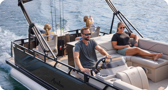 A man expertly steers a top pontoon boat brand on the water while a woman lounges beside him. Two children sit at the back, delighting in the ride. Fishing rods line the boat, and the sun blazes brightly overhead.