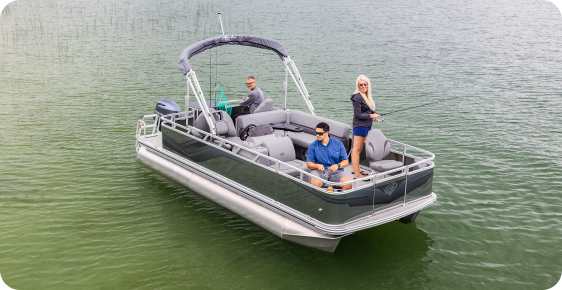 Three people are on a pontoon boat, one of the leading pontoon boat brands, enjoying a serene day on a calm lake. Two are sitting, while one stands casting a line into the surrounding green waters.