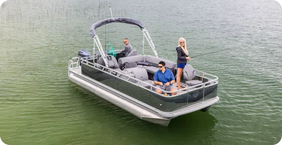 Three people are on a pontoon boat, one of the leading pontoon boat brands, enjoying a serene day on a calm lake. Two are sitting, while one stands casting a line into the surrounding green waters.