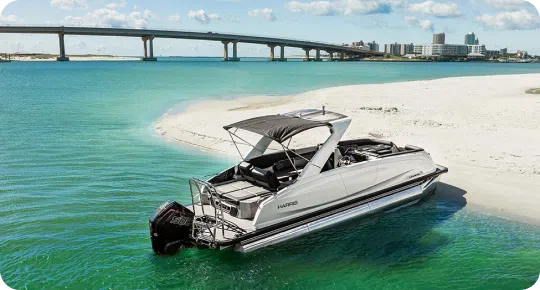 A sleek white pontoon boat with a black canopy is anchored near a sandy beach. The clear turquoise water surrounds it, with a long bridge and cityscape in the background under a clear blue sky.