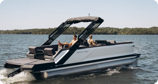 A modern pontoon boat from top brands glides across the lake, where two people relax under the sleek canopy. Lush green trees frame the scene against a clear blue sky, epitomizing leisure at its finest.