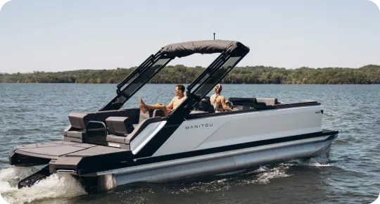 A modern pontoon boat from top brands glides across the lake, where two people relax under the sleek canopy. Lush green trees frame the scene against a clear blue sky, epitomizing leisure at its finest.