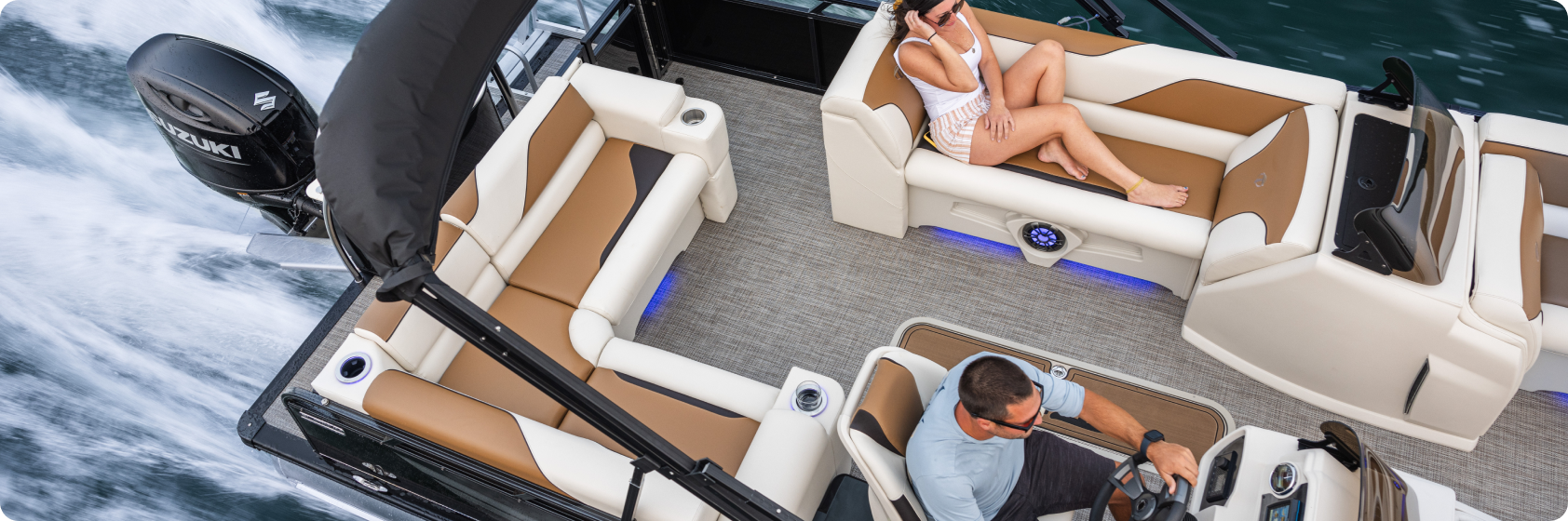 A man is steering a pontoon boat with plush beige seating, while a woman relaxes opposite him. The boat glides swiftly over the water, showcasing blue accent lighting that complements its luxurious design—a testament to top pontoon boat brands.