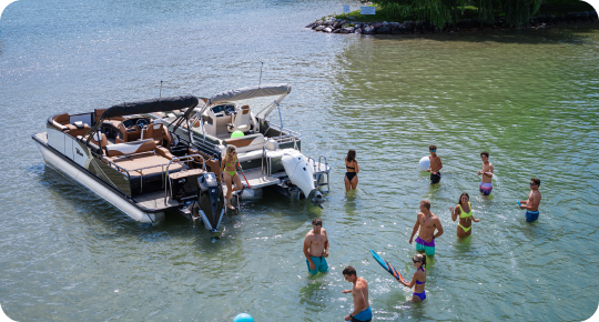 A group of people enjoy a sunny day, swimming by a pontoon boat from popular brands. The water is clear and shallow, with the shoreline close by. Everyone is in swimwear, some holding pool toys, making the most of the relaxing atmosphere by the boat.