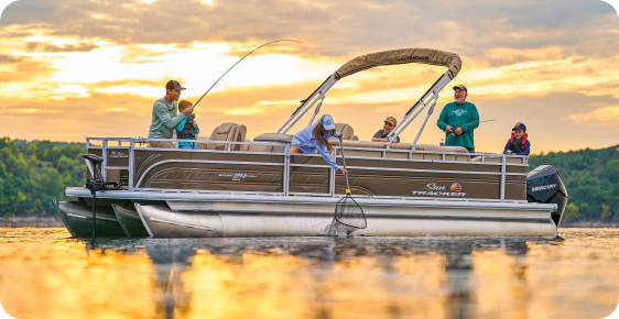 A family of six enjoys fishing on their trusty pontoon boat at sunset. One person is reeling in a fish while another uses a net. The sky is filled with vibrant clouds, and the surrounding landscape is lush and green, highlighting why top pontoon boat brands are perfect for such serene moments.