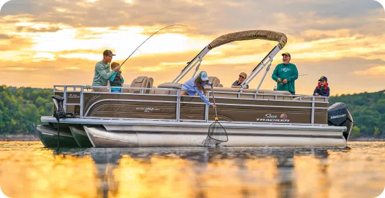 A family of six enjoys fishing on their trusty pontoon boat at sunset. One person is reeling in a fish while another uses a net. The sky is filled with vibrant clouds, and the surrounding landscape is lush and green, highlighting why top pontoon boat brands are perfect for such serene moments.