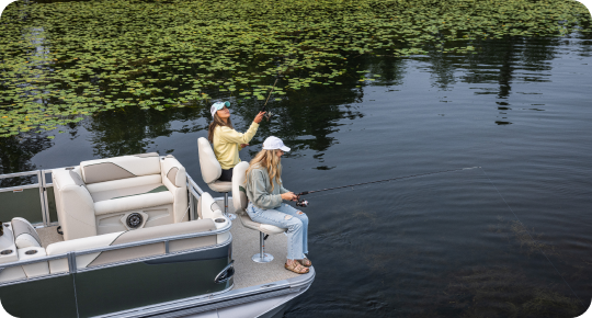 Two people are fishing from a pontoon boat on a calm, lily pad-covered lake. Both are wearing hats and casual clothing, seated as they hold fishing rods over the side. The surrounding water is tranquil, reflecting the sky—a perfect day for testing out popular pontoon boat brands.