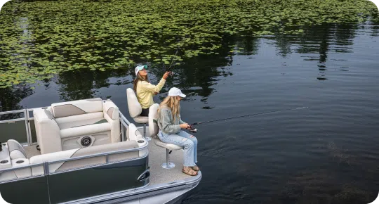 Two people are fishing from a pontoon boat on a calm, lily pad-covered lake. Both are wearing hats and casual clothing, seated as they hold fishing rods over the side. The surrounding water is tranquil, reflecting the sky—a perfect day for testing out popular pontoon boat brands.