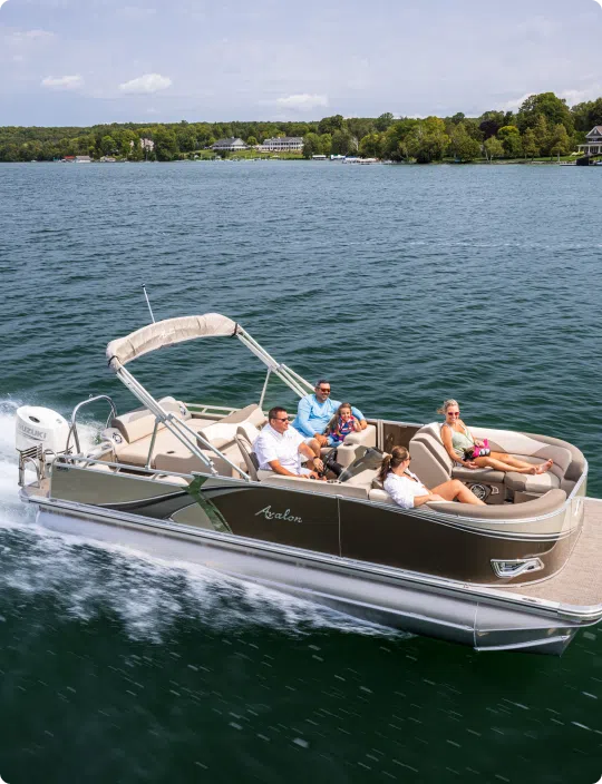 A pontoon boat with a white canopy is cruising on a lake with five people on board, showcasing one of the top pontoon boat brands. The passengers are relaxing and enjoying the sunny weather. Green hills and trees form the backdrop under a partly cloudy sky.