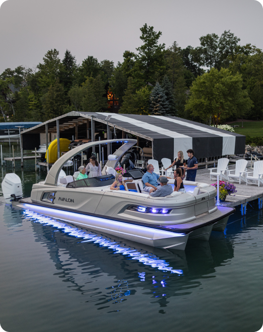 A pontoon boat from top brands, adorned with LED lights, is docked at a tranquil lakeside where several people are relaxing on board. The calm lake reflects the serene beauty of trees lining the shore, while white chairs and potted plants enhance the cozy dock ambiance.