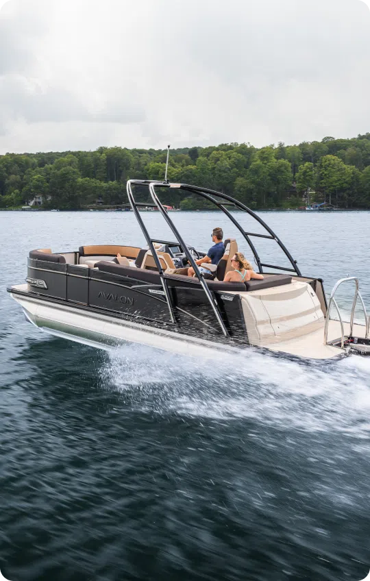 A sleek pontoon boat from top brands glides swiftly across a lake, creating a trail of white spray. A man and a woman are seated onboard, enjoying the ride. The distant shoreline is lined with dense, green trees under a cloudy sky.