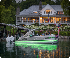 A luxurious house with large windows is illuminated in the background. In the foreground, a pontoon boat, known for its competitive pontoon prices, glows with green lights by a lush, landscaped shore. Several people are sitting on the boat, savoring the evening.