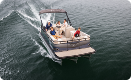 A pontoon boat with five people on board cruises across open water, embodying a picture of leisure. The group is relaxed, some seated while one steers, leaving a gentle wake behind. It's an idyllic day on the water that makes you ponder current pontoon prices for your own adventure.