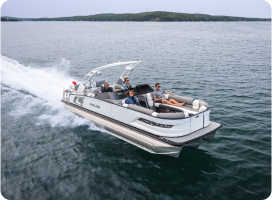 A sleek white and gray pontoon boat, known for competitive pontoon prices, speeds across a calm lake with four people onboard, enjoying the ride. The boat leaves a trail of water behind, and a dense forested shoreline is visible in the distance under a clear sky.