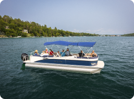 A group of people are on a pontoon boat with a blue canopy, floating on a tranquil lake. With lush trees around and houses dotting the shore, they enjoy the serene beauty while casually discussing pontoon prices under the clear sky.