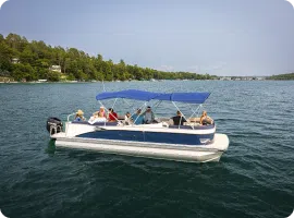 A group of people are on a pontoon boat with a blue canopy, floating on a tranquil lake. With lush trees around and houses dotting the shore, they enjoy the serene beauty while casually discussing pontoon prices under the clear sky.