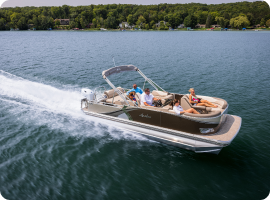 A boat speeds across a calm lake, carrying five people. The shoreline is visible in the background, lined with trees and a few houses. The sky is clear, and the water glistens in the sunlight—a perfect day to discuss pontoon prices while enjoying nature's beauty.