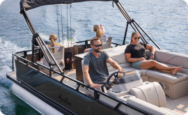 A man skillfully steers a pontoon boat on a sunny day while a woman relaxes beside him, enjoying the moment. Two children sit at the back, facing away as they take in the serene view and discuss pontoon prices. The boat glides smoothly, creating gentle waves in its wake.