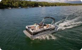 A pontoon boat glides across a large body of water under a sunny sky. Two people are relaxing and enjoying the ride, surrounded by lush green trees and distant docked boats. The wake creates a lively ripple effect, reminding them how priceless such moments are on the water.