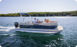 A family is enjoying a leisurely boat ride on a large lake surrounded by forested shores. The pontoon boat, with its canopy offering shade, glides across calm waters under the clear, sunny sky—a serene escape where thoughts of pontoon prices drift away with the gentle waves.