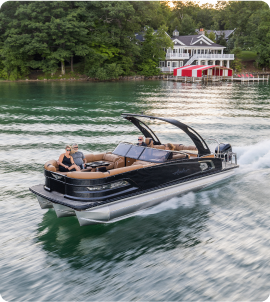 A sleek black pontoon boat with tan seating, showcasing the elegance of a new pontoon model, cruises on a calm lake. It glides past waterfront houses flanked by green trees as the sun sets, casting a warm glow on the serene scene.