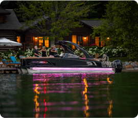 A black boat with pink lights, accompanied by a new pontoon, is docked near a wooden house by the lake. A group of people stands on the dock, and the house is surrounded by trees and illuminated with warm lights. The water reflects this enchanting scene.