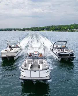 Three pontoon boats speed across a calm lake, leaving trails of white wake behind. The sky is overcast, and the shoreline is visible in the background, lined with dense green trees and a few buildings. As they glide by effortlessly, one can't help but wonder about current pontoon prices.