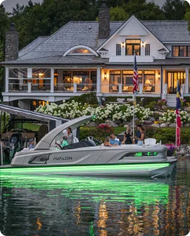 A pontoon boat with glowing green lights floats on a lake in front of a large, well-lit house, symbolizing enjoyment that surpasses mere pontoon prices. The boat carries a group of people and features flags, while the house boasts a stone chimney and a lawn adorned with blooming hydrangeas.