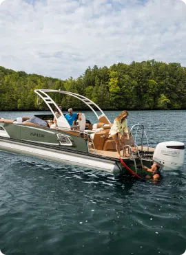 Three friends enjoy a perfect day on the water: one steering the boat, another standing, and one swimming near the back. Against a backdrop of lush green forests and a partly cloudy sky, they discuss pontoon prices while savoring their peaceful escape.