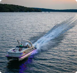 A motorboat with colorful lights glides across a vast, calm lake during sunset, reminiscent of pontoon adventures. Two people are aboard, enjoying the ride. The sky is a soft gradient of warm colors, and the distant shoreline is lined with trees.